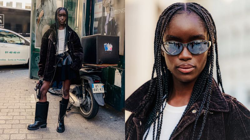 A fashionable woman with braided hair poses on a city street in a dark jacket, black skirt, and boots, with a motorcycle in the background.