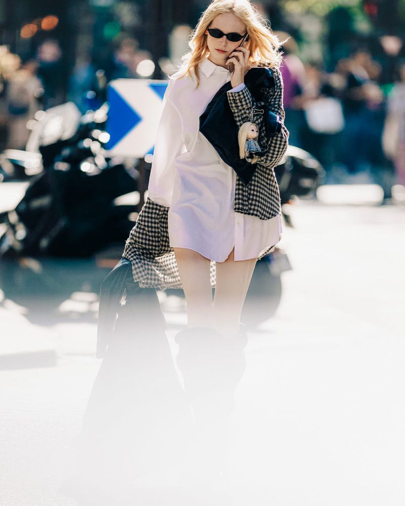 A blonde woman in a white shirt and houndstooth jacket is talking on her phone outside, captured in an eye-level outdoor shot.