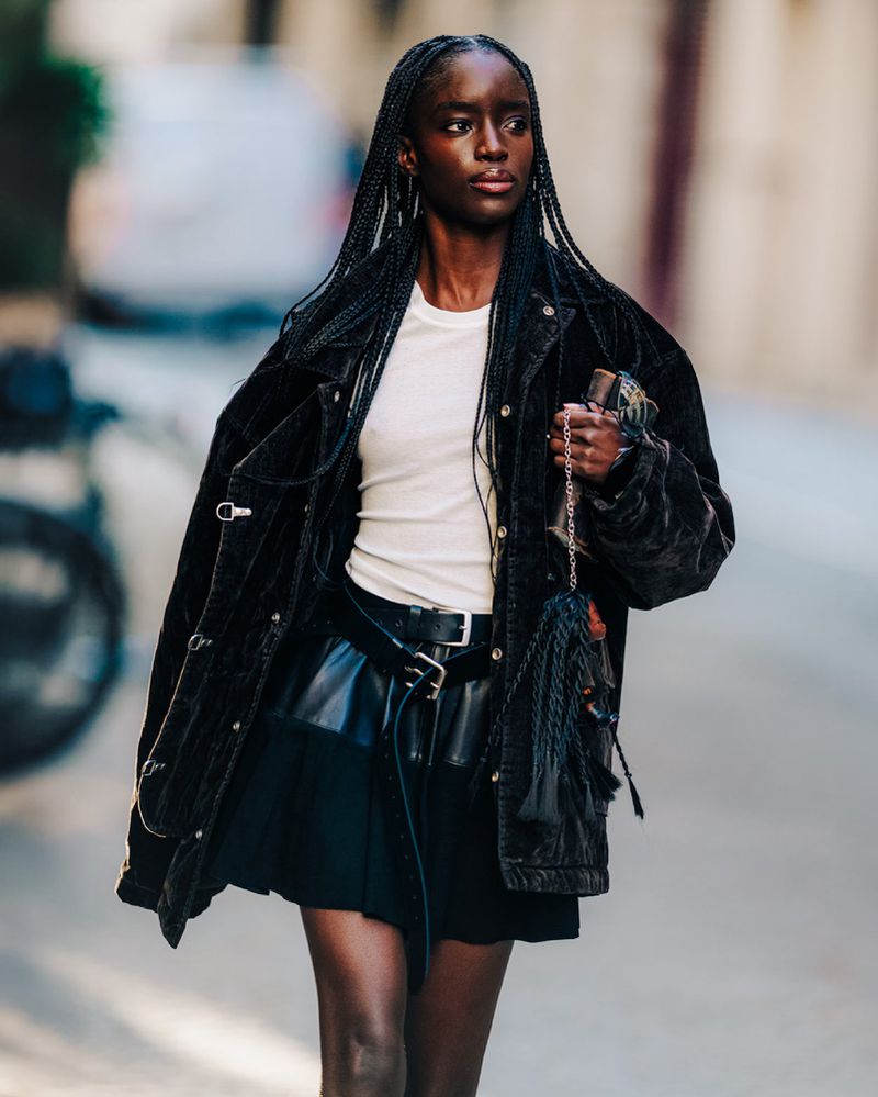 An adult Black woman poses on a city street wearing a black jacket and skirt, paired with a cream-colored top, jewelry, and a black handbag.