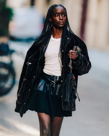 An adult Black woman poses on a city street wearing a black jacket and skirt, paired with a cream-colored top, jewelry, and a black handbag.