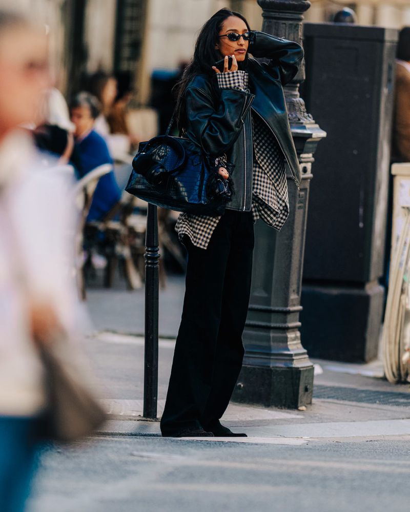 A woman in a black leather jacket and sunglasses stands on a city street with a black purse and patterned shirt, looking fashionable.