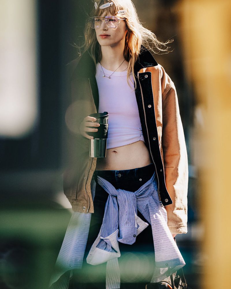 A young adult female stands outdoors holding a travel mug, wearing a crop top, jacket, jeans, and glasses in a lifestyle shot.