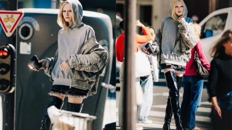 Blonde model wearing an oversized grey hoodie, bomber jacket, and black patent thigh-high boots walking on a city street.