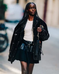A Black woman with long braided hair walks outdoors wearing an oversized black jacket, white t-shirt, and black pleated mini skirt.