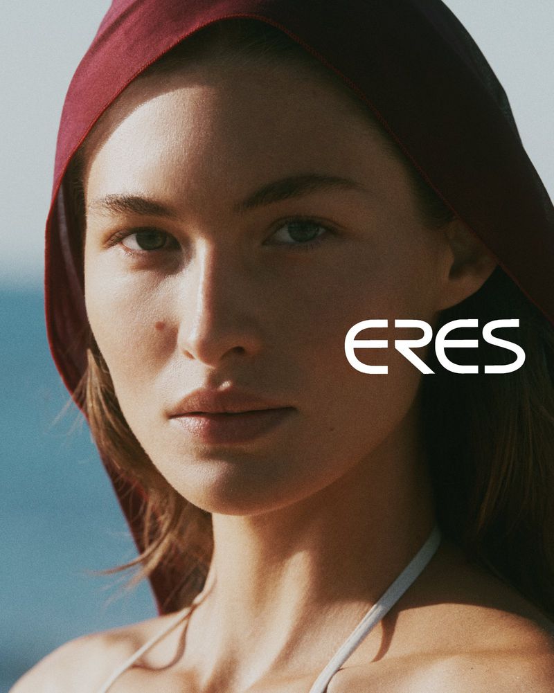 Close-up studio shot of a woman wearing a red hood and white bathing suit top, with a beach and blue sky in the background.