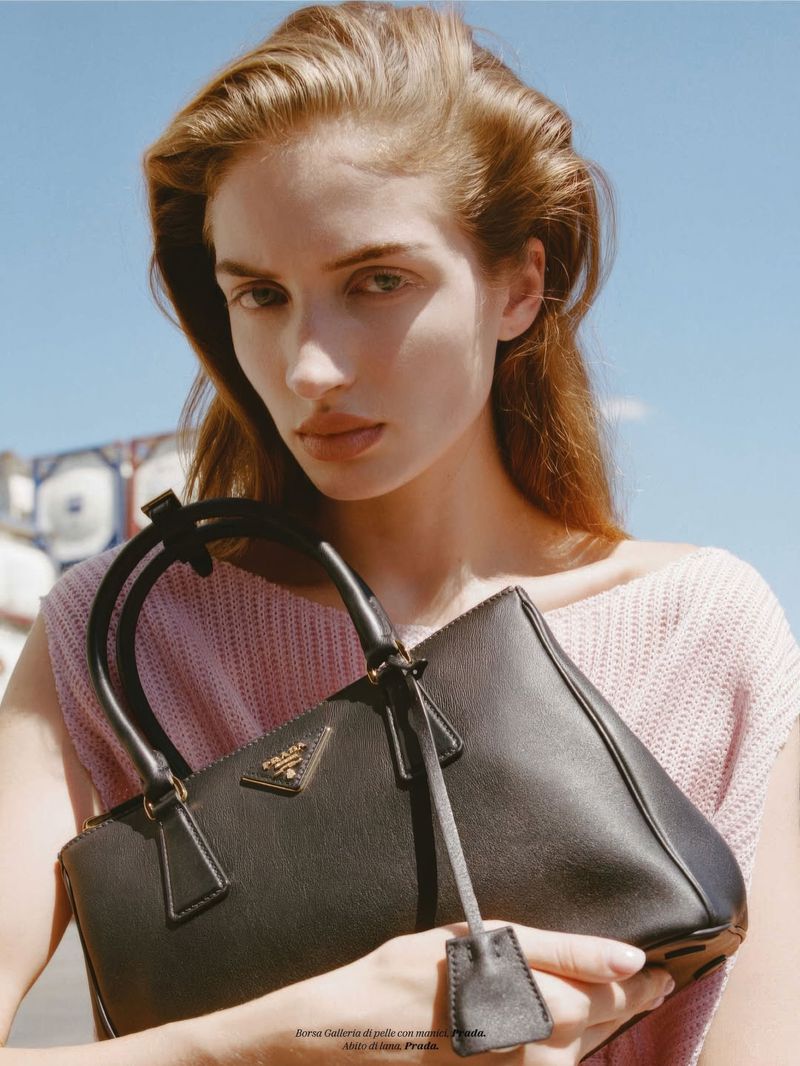 Portrait of a young woman with long red hair and a black Prada handbag, posed against a bright blue sky in an outdoor setting.