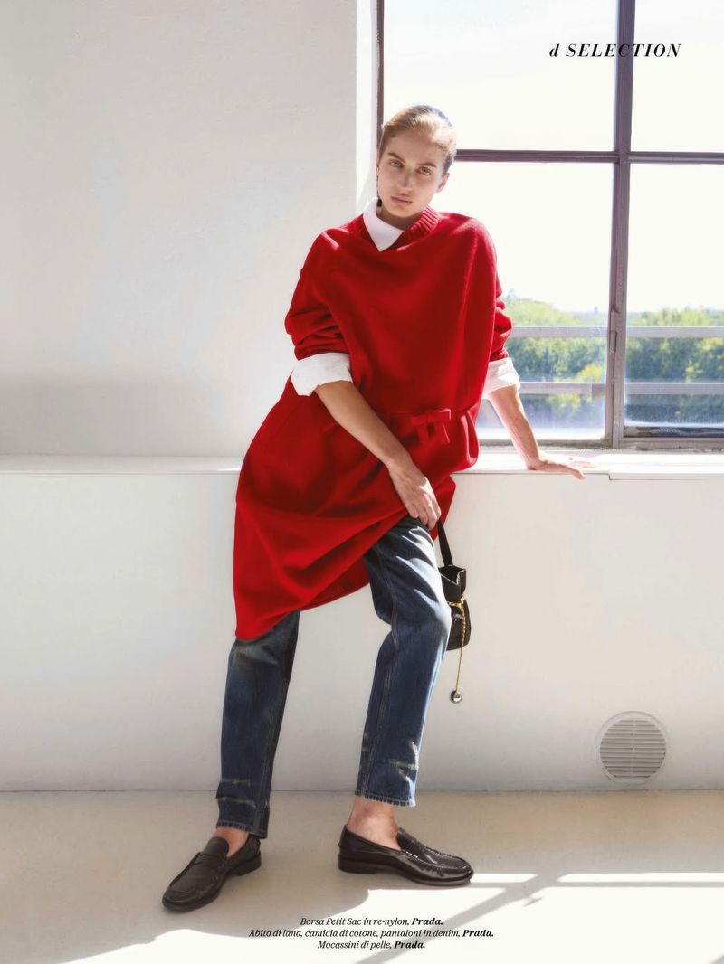 A fashionable young woman in a red sweater dress, jeans, and loafers poses indoors near a window with muted outdoor light.