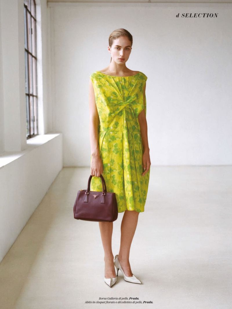 A woman poses in a yellow floral dress with a handbag, set against a bright studio backdrop with a window. Fashion editorial.