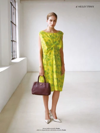 A woman poses in a yellow floral dress with a handbag, set against a bright studio backdrop with a window. Fashion editorial.