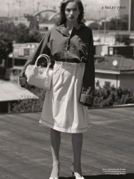 Model standing on a rooftop holding a light handbag wearing a dark buttoned top and a light pleated skirt against an urban background