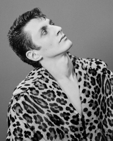 Black and white portrait of a young adult male wearing a leopard print jacket, looking up and to the side against a grey background.