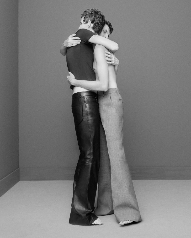 Black and white studio shot of two adults hugging, one wearing black leather pants and a t-shirt, the other in flared trousers.