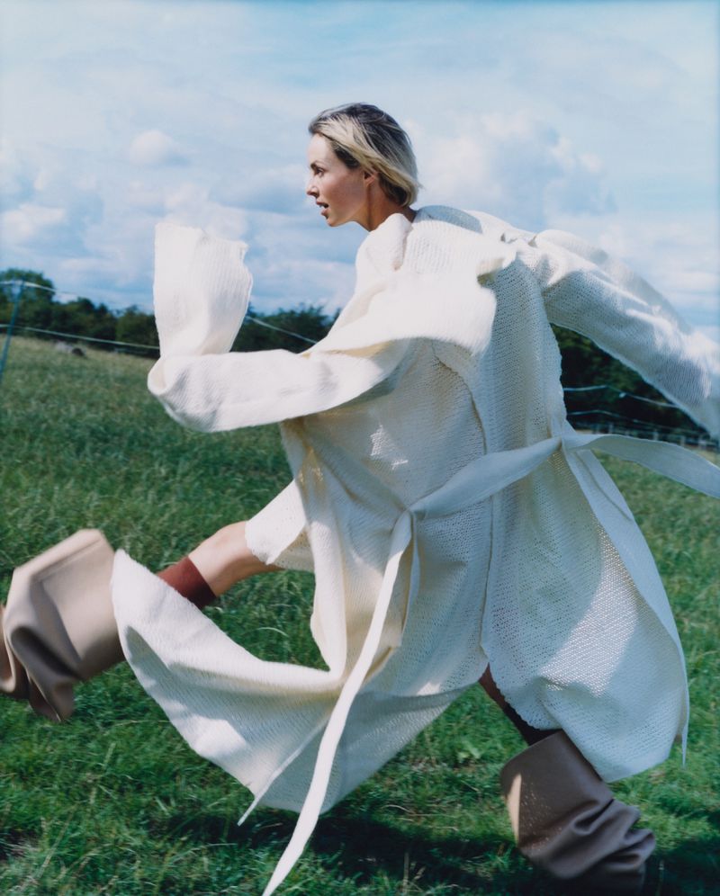 Woman runs through a grassy field wearing a cream-colored coat and tan boots against a backdrop of trees and a cloudy sky.
