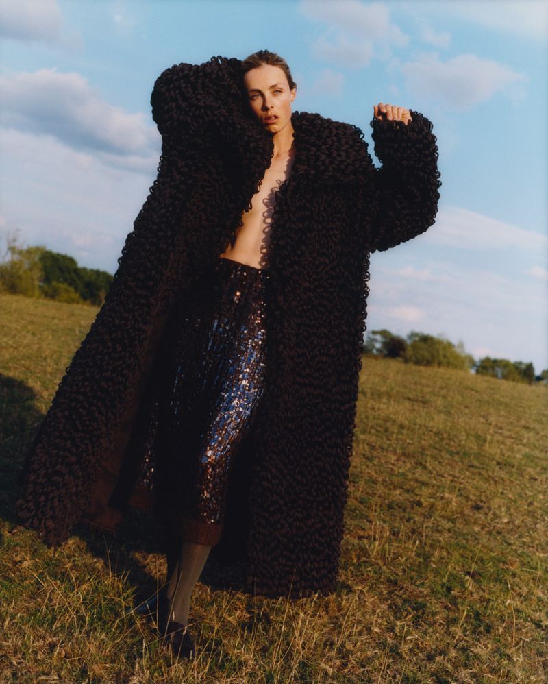 Woman in dramatic coat and sequined skirt poses in a grassy field under a blue sky with scattered clouds, fashion outdoor setting.