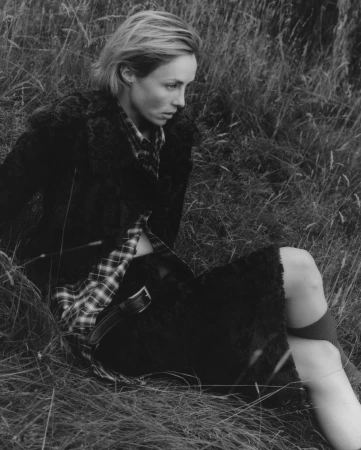 Blonde woman in coat and flannel sitting in tall grass in black and white. Fashion shot in a rural landscape. Model posing outdoors.