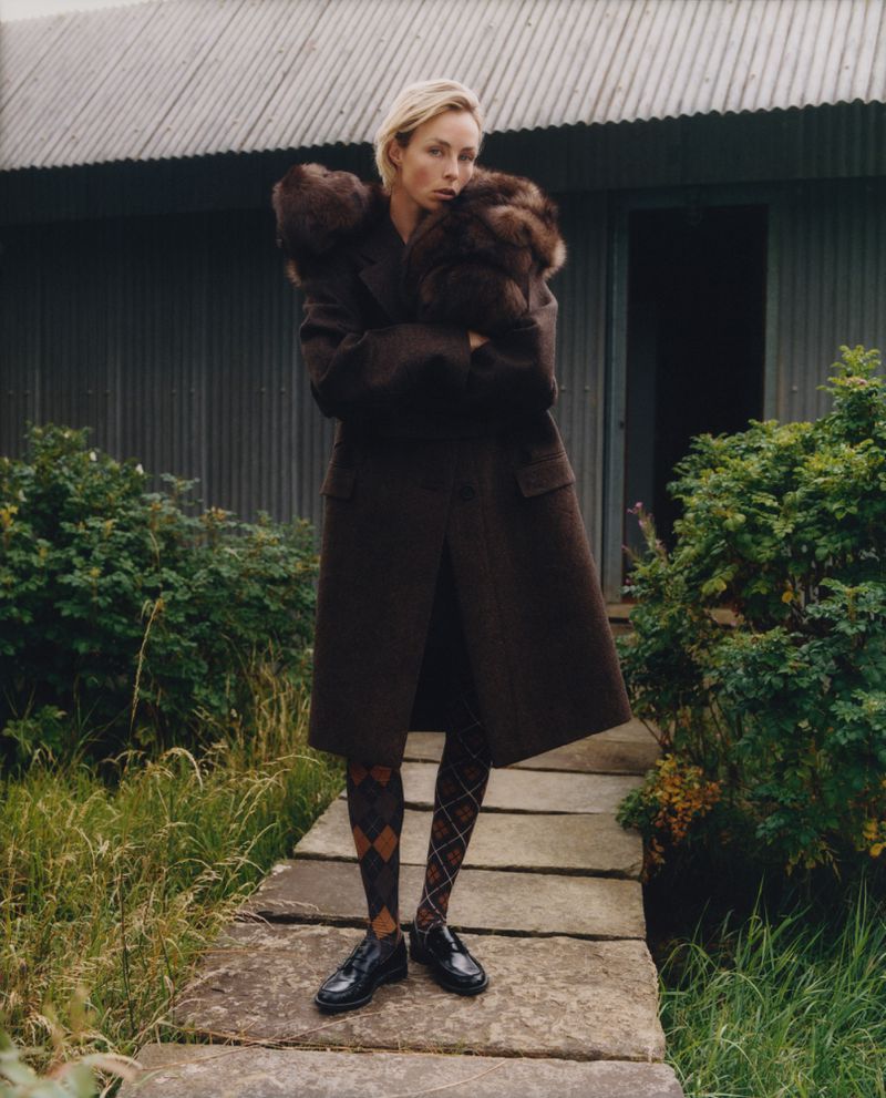 Blonde model poses in brown fur-trimmed coat and patterned tights on a stone path with a corrugated building behind her outdoors.
