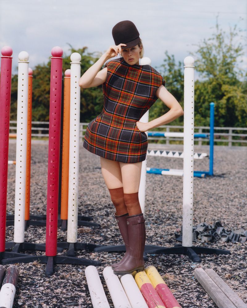 Woman poses outdoors wearing a plaid minidress, brown riding boots, and a cap, surrounded by riding arena equipment.
