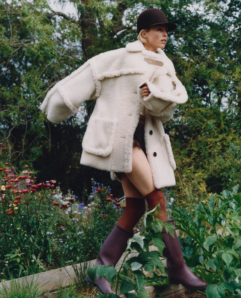 A fair-skinned adult woman with short blonde hair and a brown hat walks through a flower garden wearing a cream-colored shearling coat and brown boots.