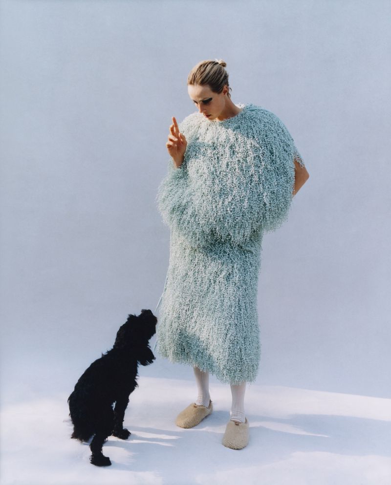 Woman in a light blue shaggy outfit stands in a bright setting gesturing to a black dog; studio lighting and neutral backdrop.