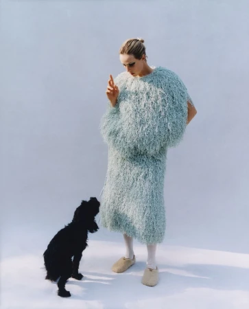 Woman in a light blue shaggy outfit stands in a bright setting gesturing to a black dog; studio lighting and neutral backdrop.