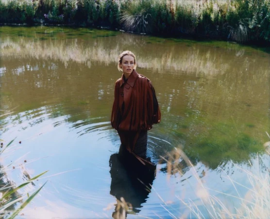 Woman in a red coat stands knee-deep in a pond reflecting the sky. Green reeds line the background in an outdoor daytime setting.