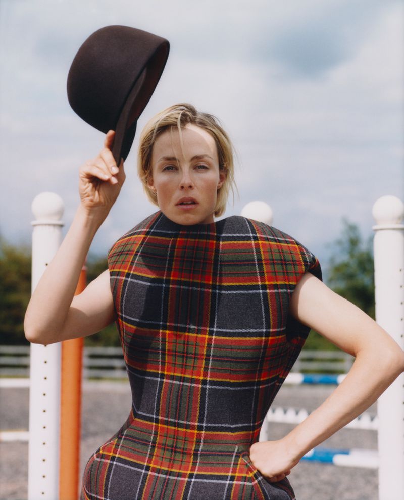 Adult woman in plaid dress with bowler hat in outdoor fashion shot with geometric fence, blue sky, and green trees.