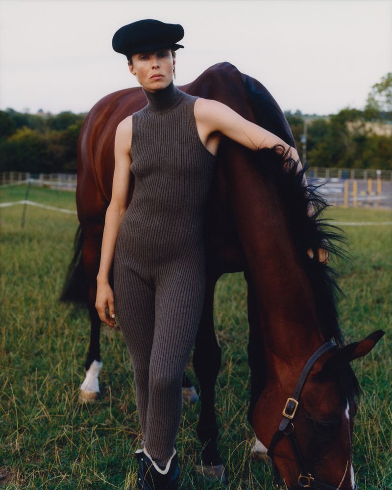 Woman in a sleeveless catsuit and cap stands next to a brown horse in a grassy field with trees and a cloudy sky in the background.