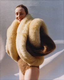 Model Edie Campbell wearing a textured brown bodysuit and a large voluminous cream fur wrap against a neutral textured background.