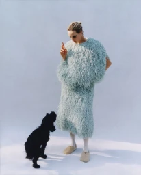 Model Edie Campbell in a shaggy light mint green dress looking down at a small black dog against a white background.