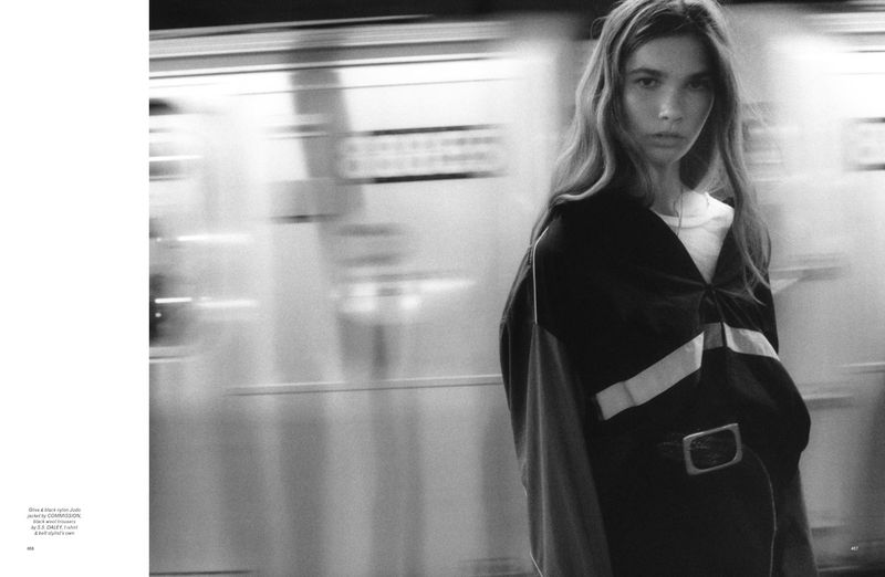 Black-and-white studio shot of an adult female model wearing an oversized black jacket, standing in front of an out-of-focus subway train.