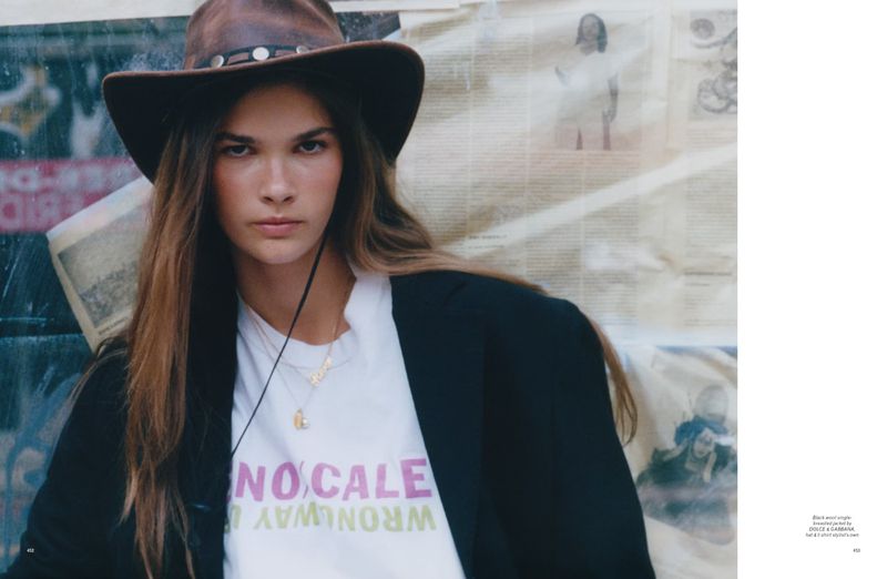 A woman with long hair wearing a cowboy hat and a black jacket poses in front of a newspaper-covered background, looking at the camera.