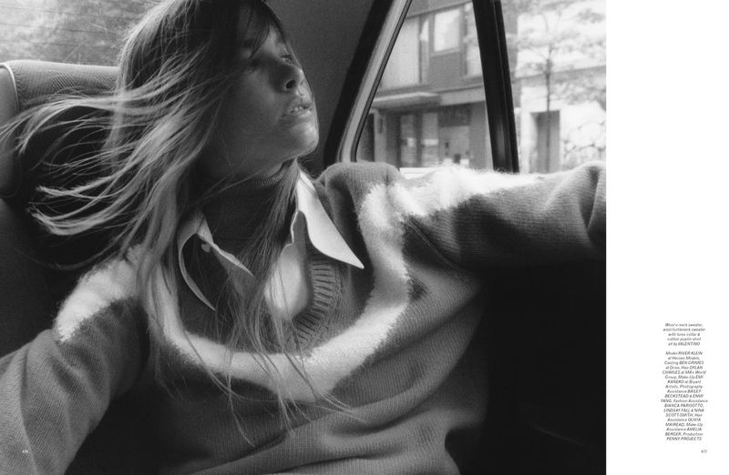 Monochrome indoor portrait of a woman with flowing hair in a patterned sweater sitting in a car next to a window.