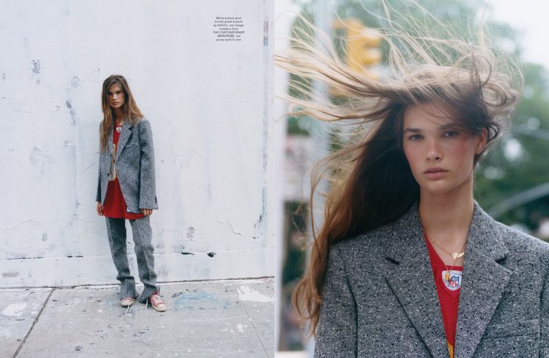 Fashion editorial photo featuring a young woman in a gray suit and red top, posing on a city street with blowing hair and a serious expression.