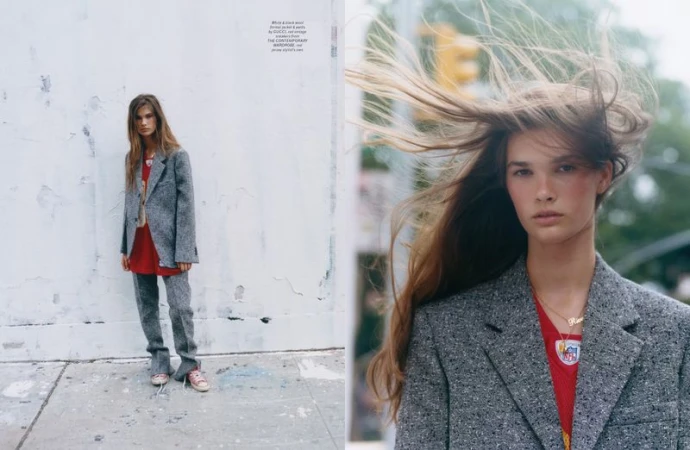 Fashion editorial photo featuring a young woman in a gray suit and red top, posing on a city street with blowing hair and a serious expression.