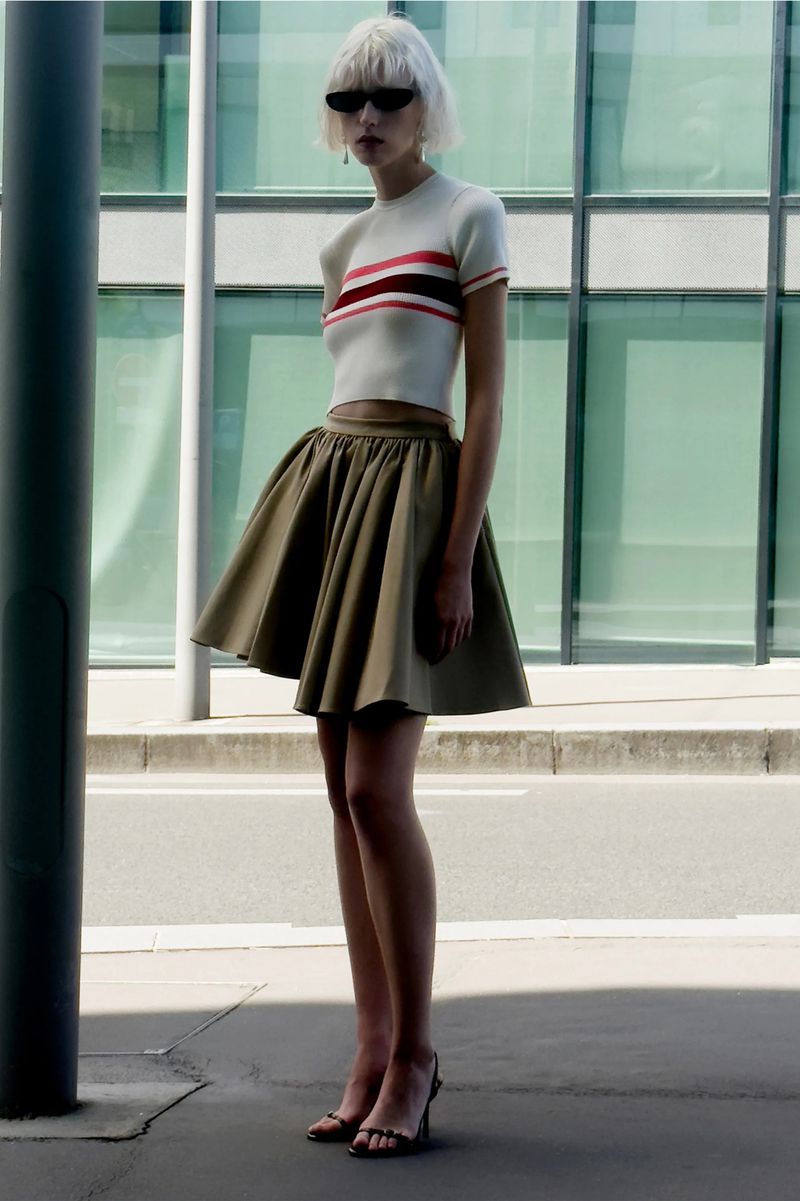 Fashion shot of a slender woman with a blonde bob, wearing sunglasses, a short white top, and a flared olive green skirt, posing outdoors.