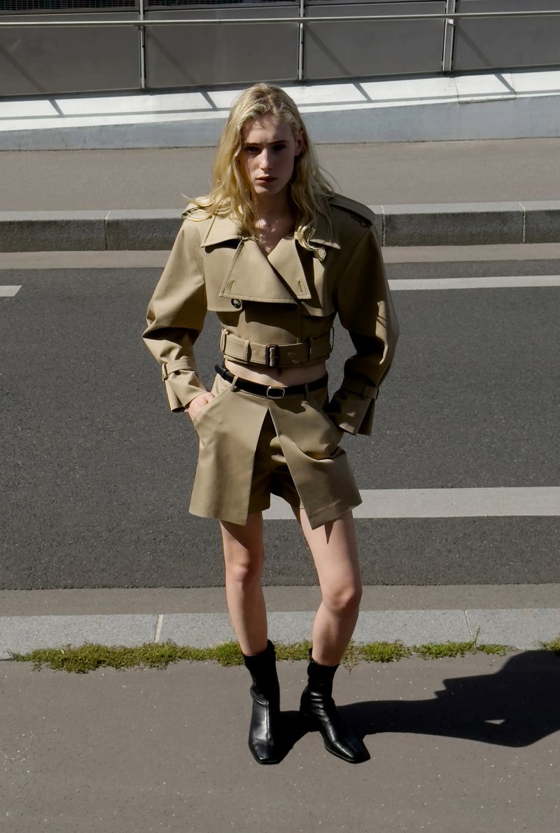 A blonde female model stands on a city street wearing a cropped tan trench coat, matching skirt, black belt, and black boots.