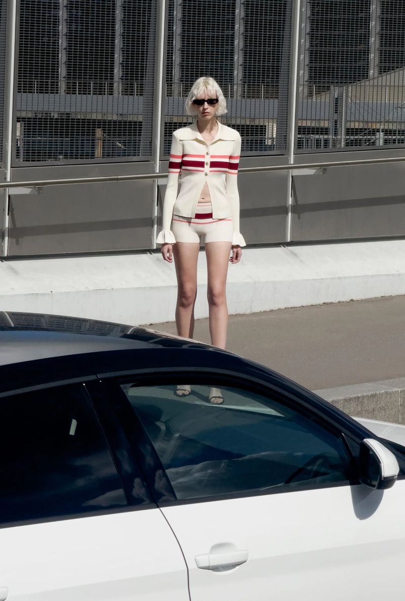 A slender woman with blonde hair, wearing sunglasses and a knit outfit, stands outdoors by a modern white car in daylight.