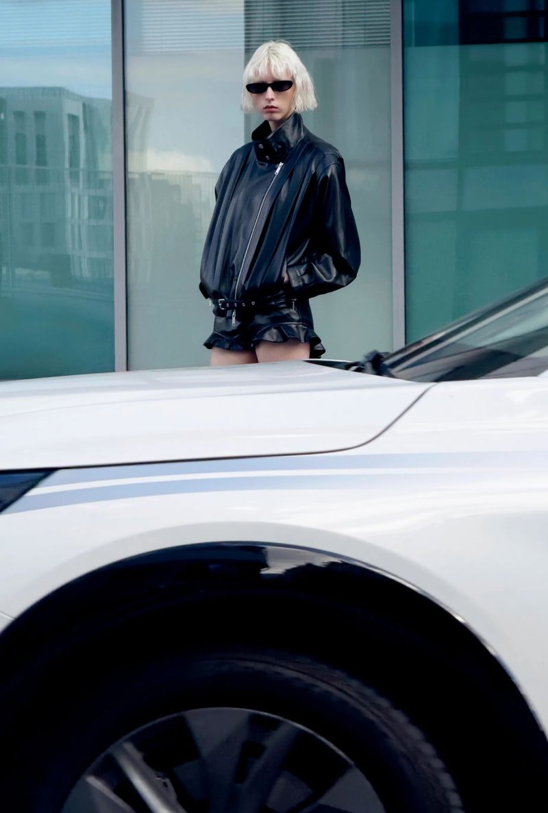 A woman with blonde hair wearing a black leather jacket and shorts poses next to a white car in front of a glass building.