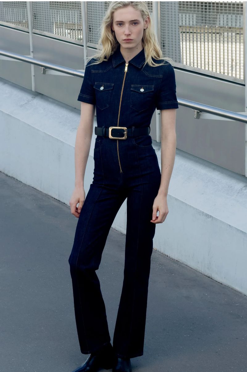 Woman in dark denim jumpsuit with gold zipper and black belt stands on gray pavement near a railing, casting a neutral expression.