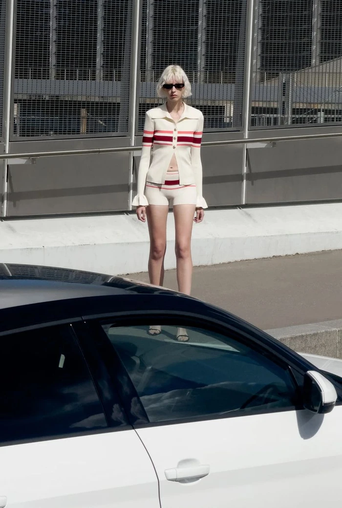 Model with blonde bob and sunglasses wearing a striped cream knit top and shorts set behind a white car in an urban setting.