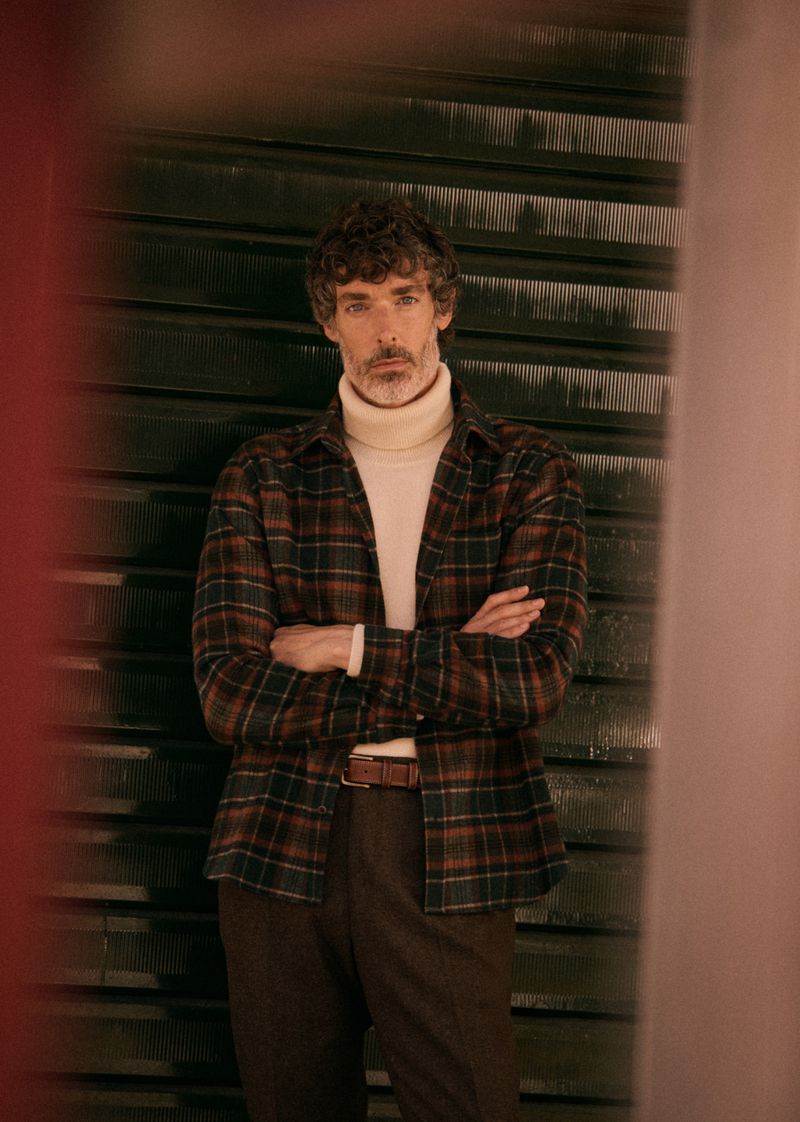 Adult male with curly hair, in a plaid shirt and cream turtleneck, stands with arms crossed against a textured metal background.