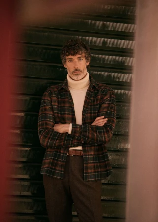 Adult male with curly hair, in a plaid shirt and cream turtleneck, stands with arms crossed against a textured metal background.