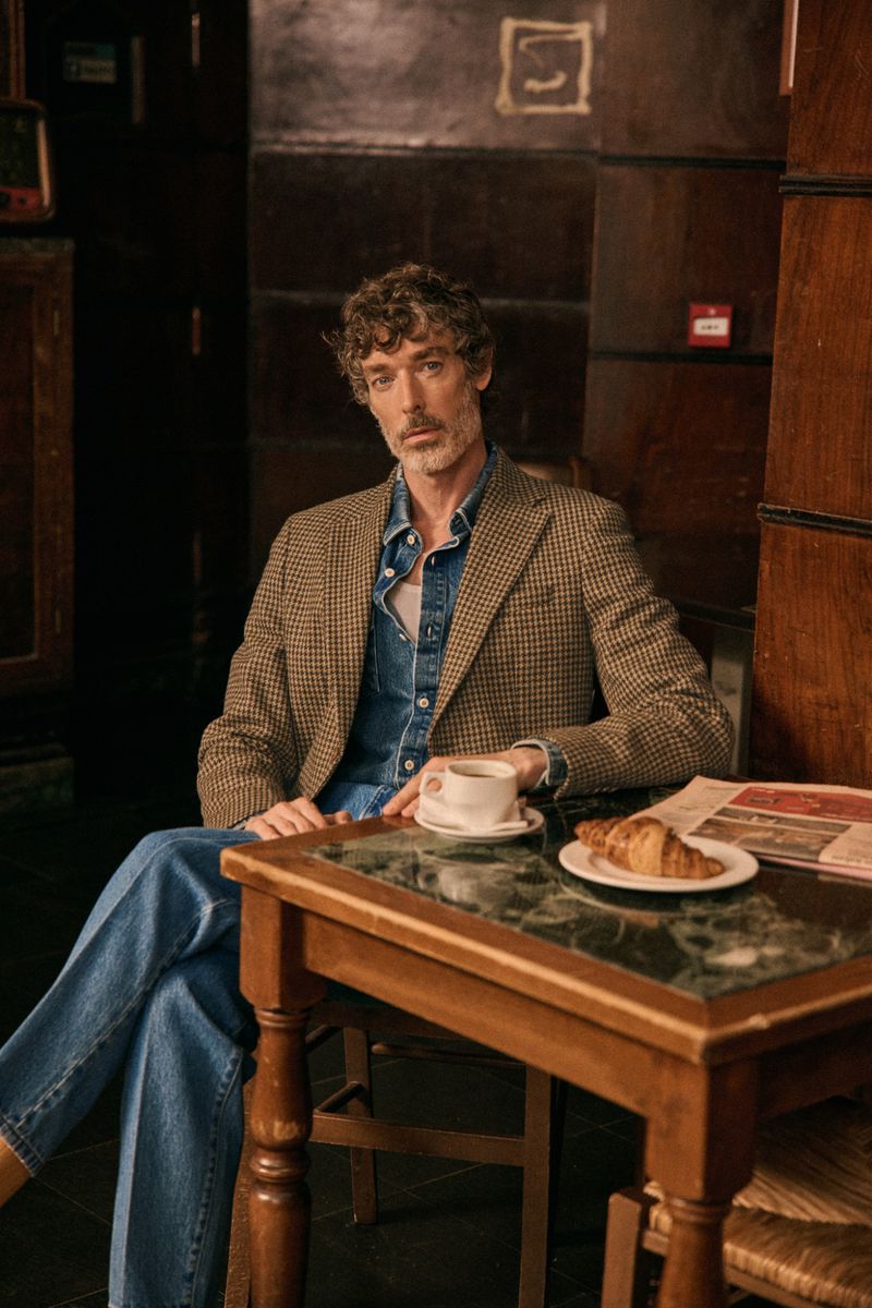 A man in a blazer and jeans sits at a cafe table with a coffee and croissant in a warm-toned indoor setting, styled for fashion editorial.