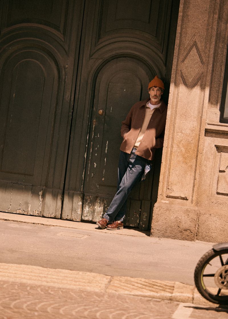 Adult male leaning against a stone building wearing a jacket and beanie, with a partial view of a bicycle wheel on the sidewalk.