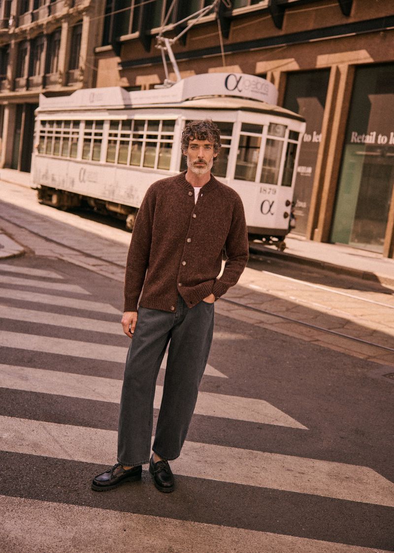 Adult male poses in brown cardigan and grey trousers on a city street with tram, showcasing fashion and urban style in natural light.