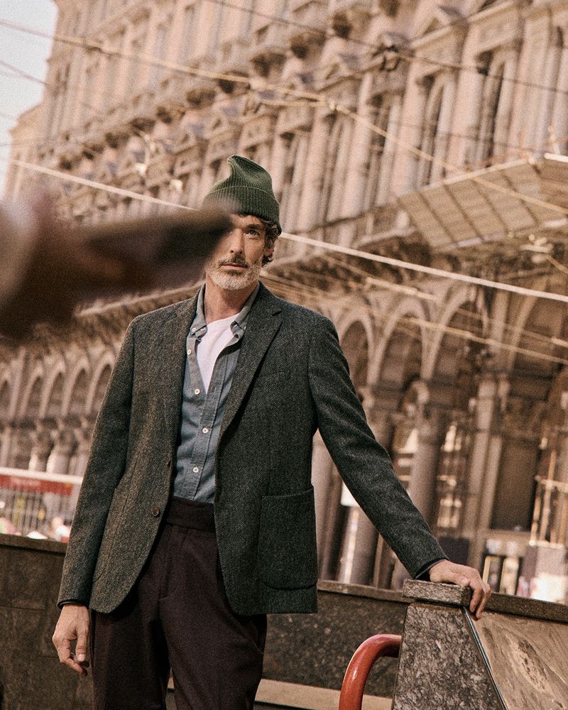 Male fashion model in a blazer and beanie poses in an urban setting, featuring classic architecture. Street style with a European flair.