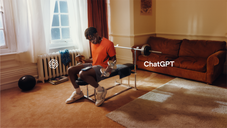 Young man in an orange shirt works out indoors with dumbbells on a weight bench, with a sofa and radiator visible in the background.