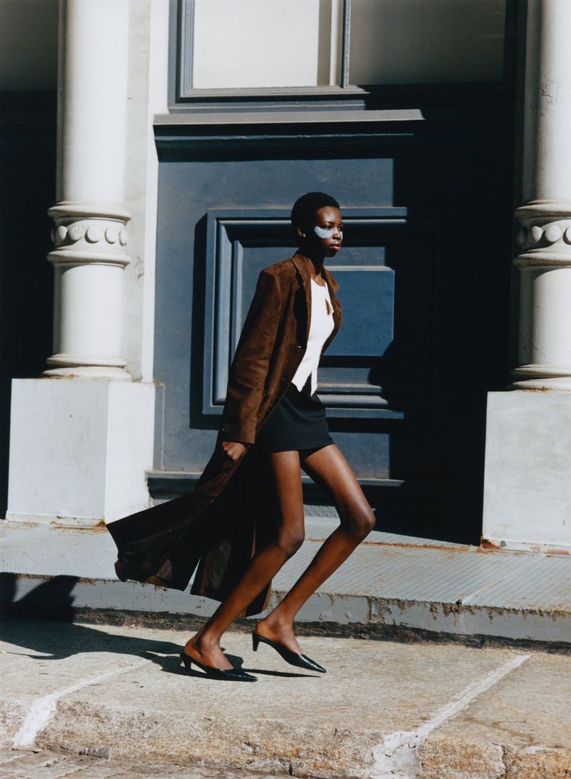 A fashionably dressed woman in a brown coat and black mini-skirt walks down stone steps in front of a grey building with white columns.
