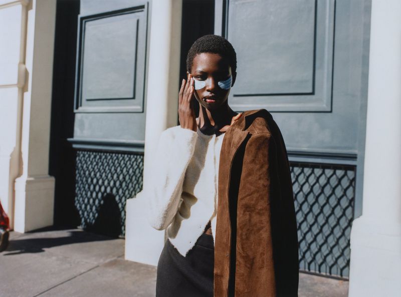 A model with eye patches poses outside wearing a white sweater and brown jacket, standing on a city street with a large blue door.