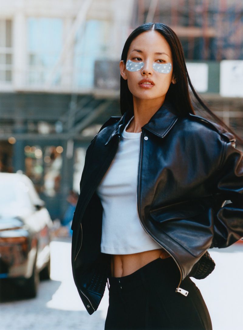 Asian woman in black leather jacket and eye patches poses on urban street with city buildings and car in the background.